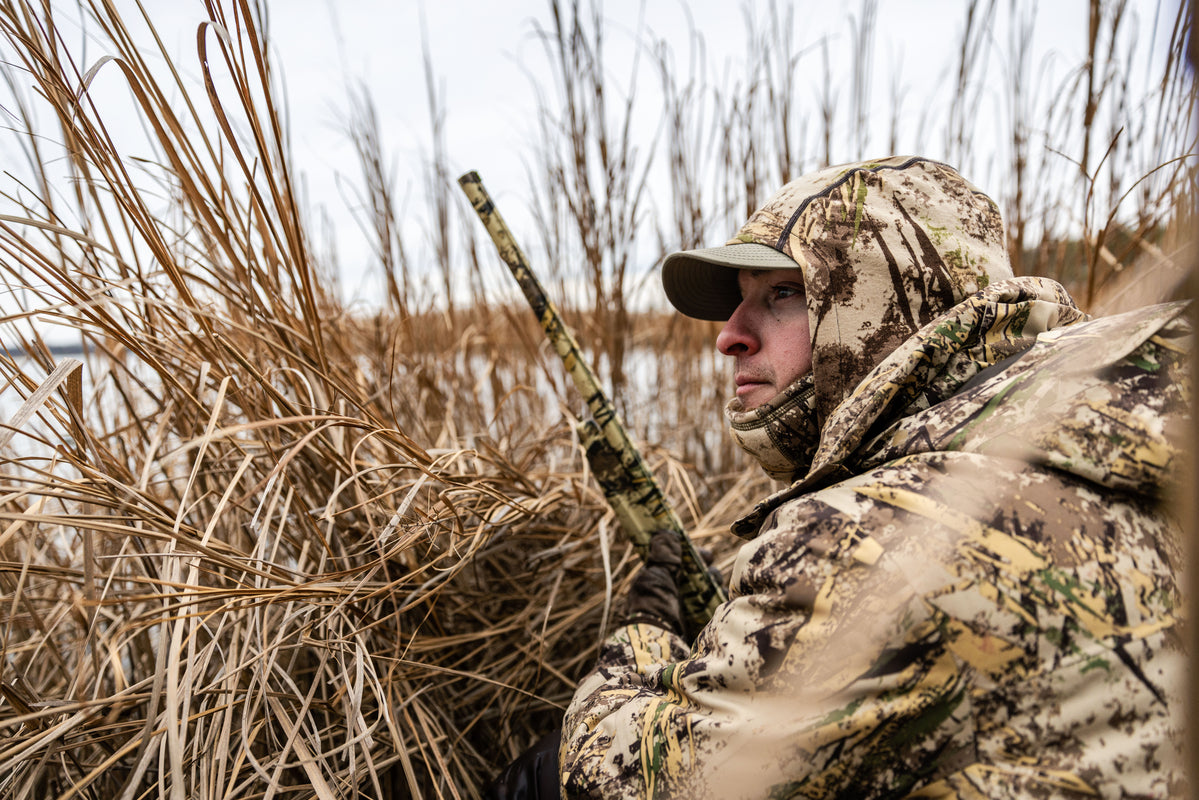 Person in camouflage clothing holding a rifle, standing in tall grass with a body of water and trees in the background.