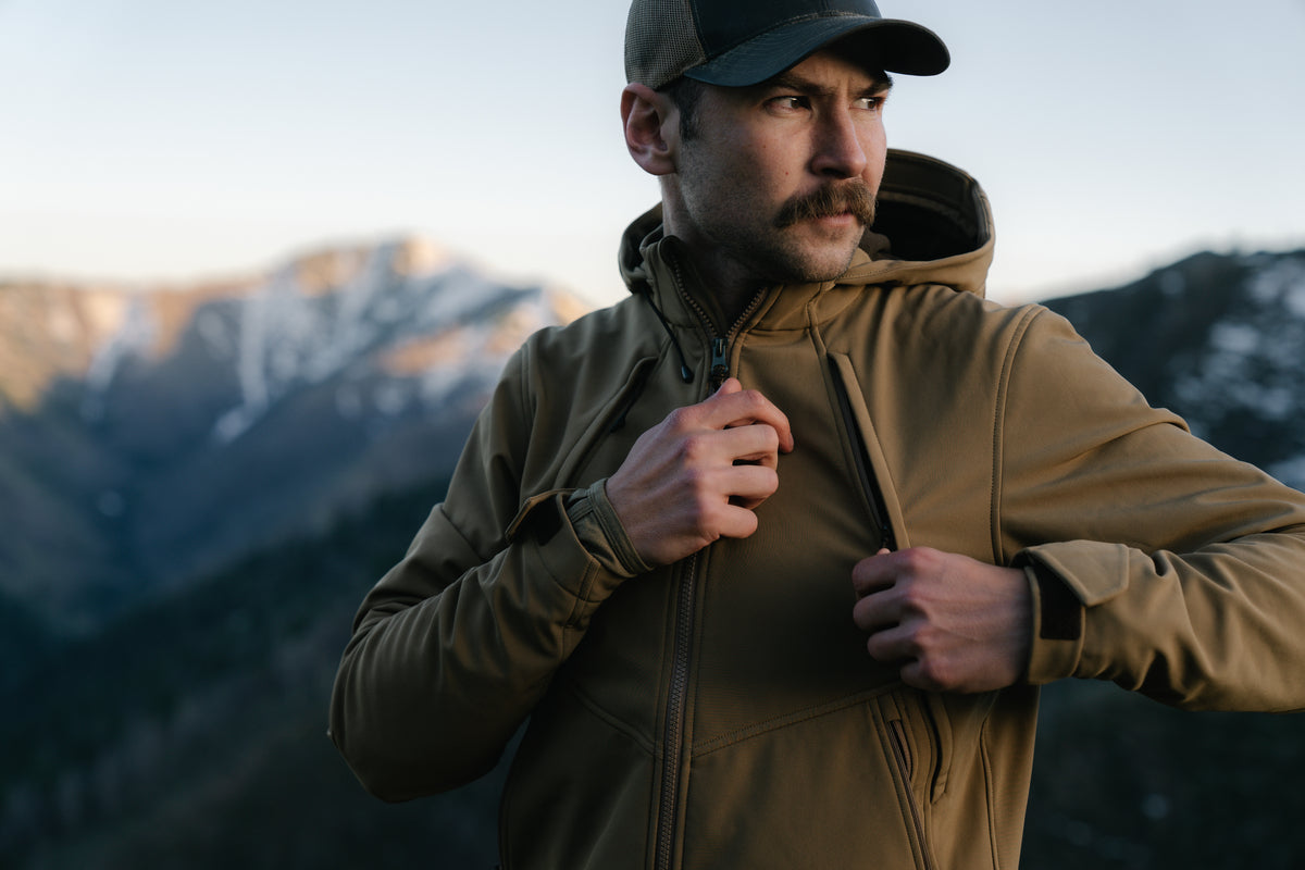 Man in a tan jacket and cap standing in a mountainous landscape