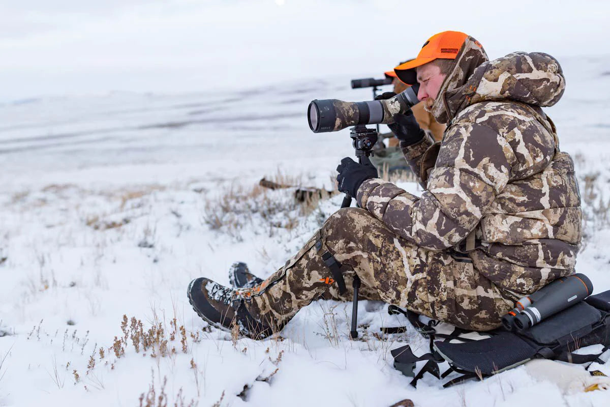Person in camouflage gear and orange cap using a rifle with scope in a snowy field