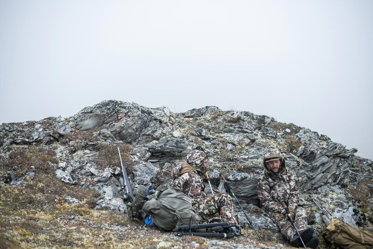 Two hunters in camouflage gear sitting on a rocky hillside with rifles.